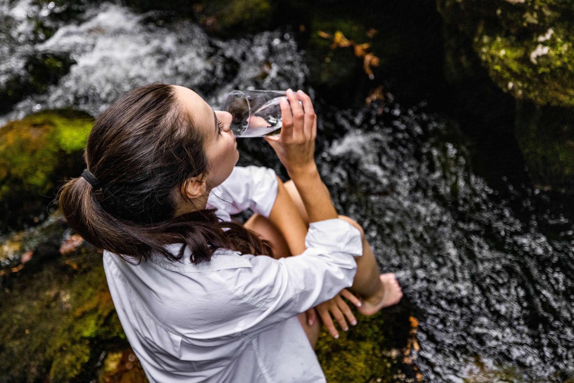 Woman drinking water while sitting on rock by river in forest